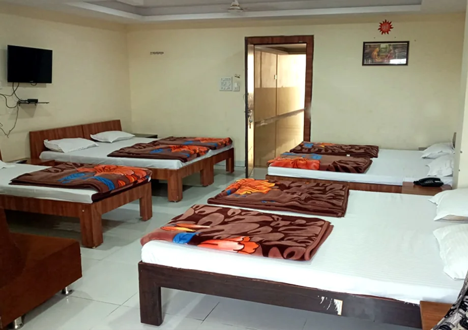 Seven-bed dormitory hall with blankets, pillows and wall-mounted TV at Narasinha Mehta Dham Girnar
